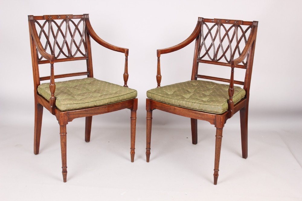 A pair of antique George III mahogany elbow chairs featuring openwork lattice backs, swept armrests, turned front legs, and green seat cushions.