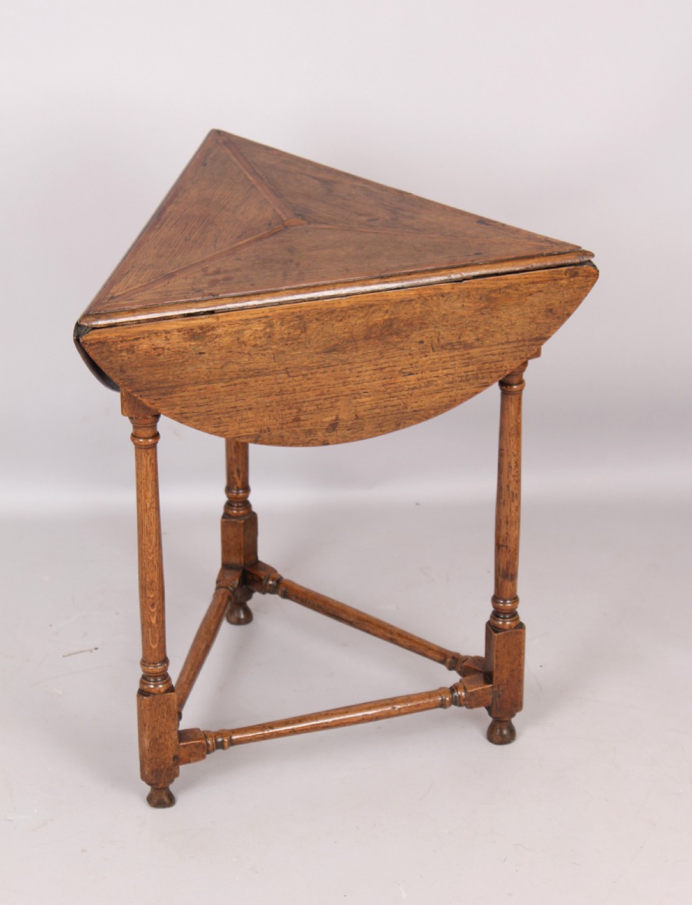 An early 18th-century oak cricket table featuring a triangular top with drop leaves, turned legs, and a triangular stretcher base.