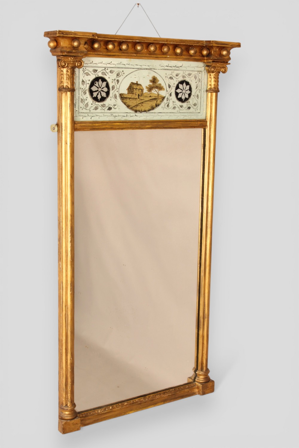 A vertical Regency giltwood wall mirror with fluted side columns, a breakfront cornice with spherules, and a reverse-painted glass panel depicting a cottage landscape above the main glass plate.