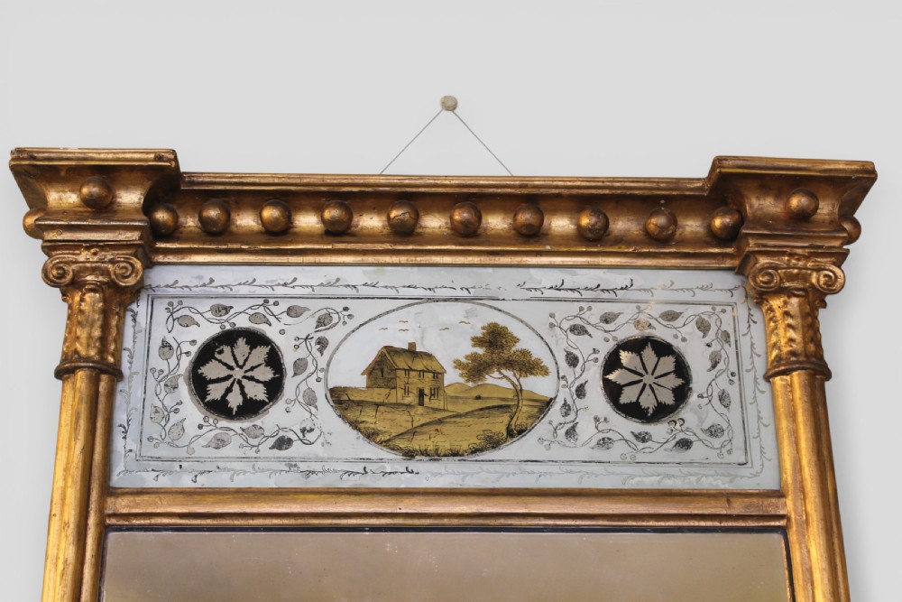A vertical Regency giltwood wall mirror with fluted side columns, a breakfront cornice with spherules, and a reverse-painted glass panel depicting a cottage landscape above the main glass plate. View II