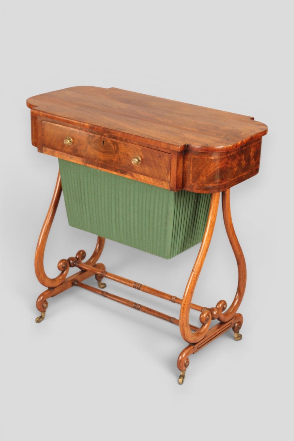 A Regency mahogany work table featuring a single drawer above a pleated green fabric basket, supported by lyre-shaped ends and a double stretcher with castors.