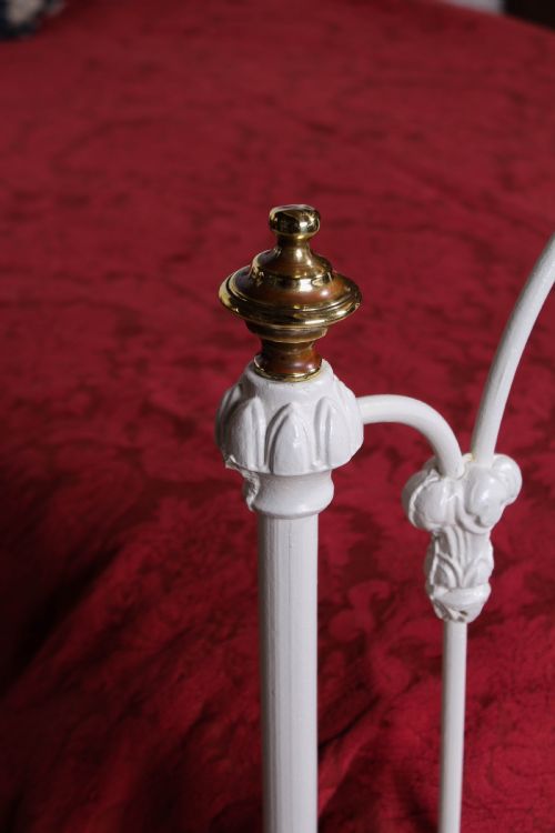 fig 1. A Victorian white painted wrought iron single bed frame featuring ornate scrollwork, floral metal castings, turned brass finials, and castors.