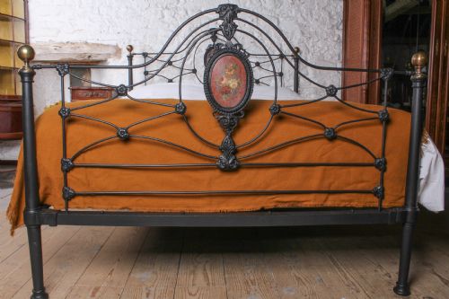 fig 1. A Victorian black-painted cast iron bedstead featuring brass finials, arched lattice scrollwork, and oval floral enamel medallions on the headboard and footboard.