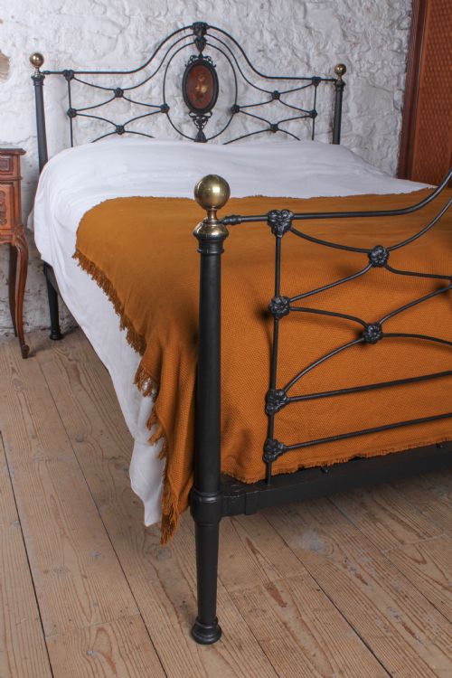 fig 5. A Victorian black-painted cast iron bedstead featuring brass finials, arched lattice scrollwork, and oval floral enamel medallions on the headboard and footboard.