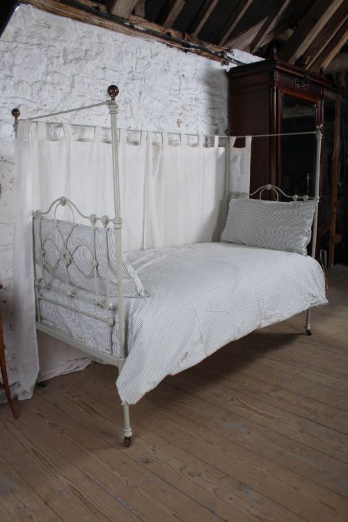 fig 1. A painted single iron four-poster bed frame featuring decorative central scrollwork, brass top finials, and lightweight drapery.