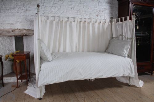 fig 3. A painted single iron four-poster bed frame featuring decorative central scrollwork, brass top finials, and lightweight drapery.