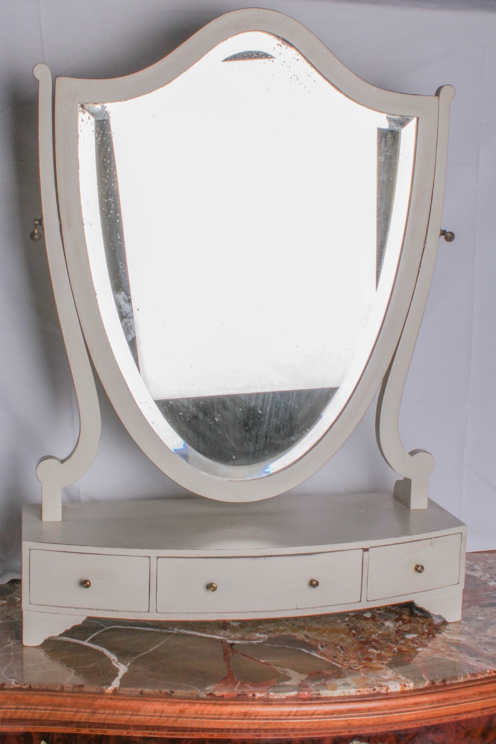 A painted off-white shield-shaped dressing mirror with a swivel glass frame set above a curved base containing three small drawers with brass knobs.