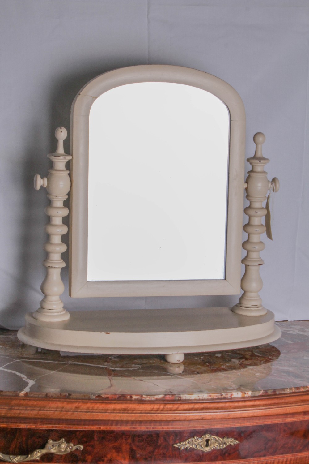 An off-white painted Victorian toilet mirror featuring an arched glass frame supported by two turned upright posts on a plinth base.
