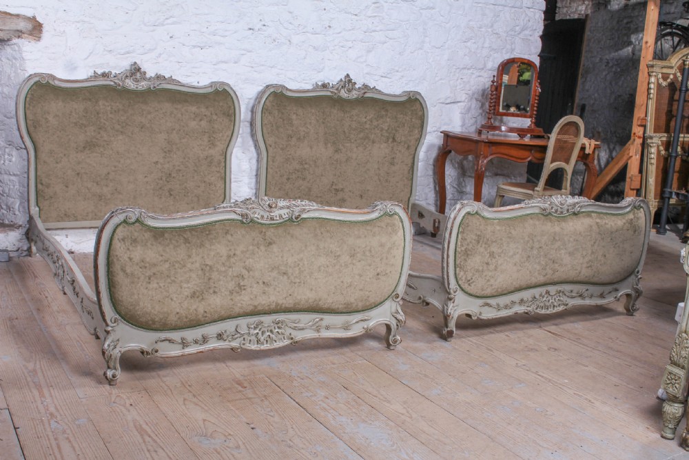 A pair of French Louis XV style single bed frames with carved grey painted timber and olive green velvet upholstered panels.