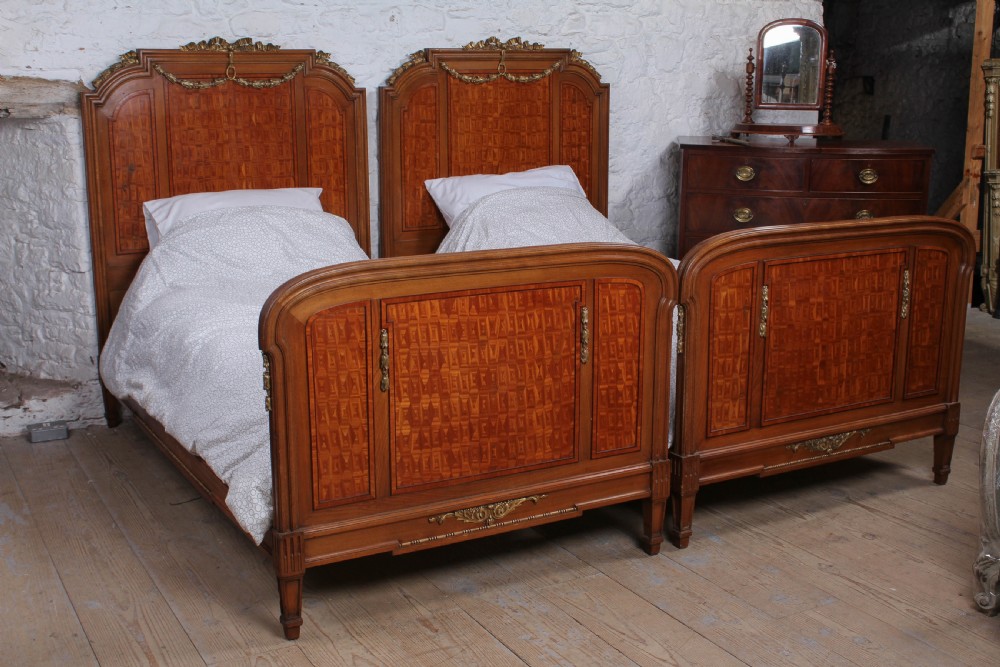 A pair of antique French fruitwood single beds featuring geometric parquetry inlay and decorative gilt bronze mounts on the headboards and footboards.