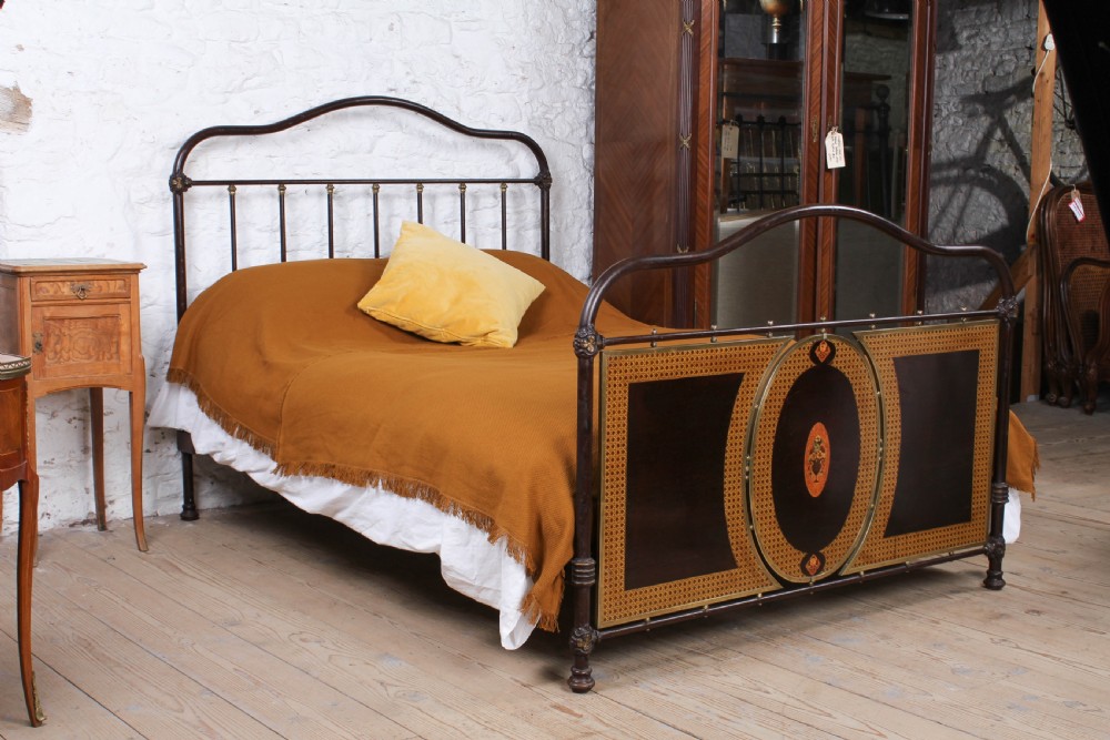 An antique European double bed featuring a dark iron frame, brass top rails, vertical spindles on the headboard, and decorative painted panel inserts on the footboard.