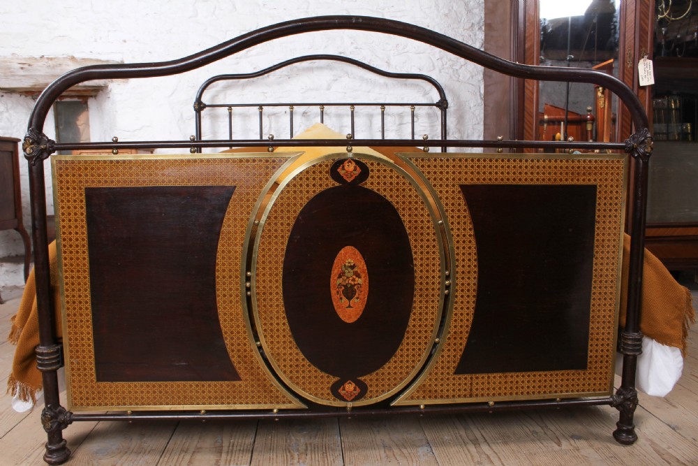 An antique European double bed featuring a dark iron frame, brass top rails, vertical spindles on the headboard, and decorative painted panel inserts on the footboard. View II