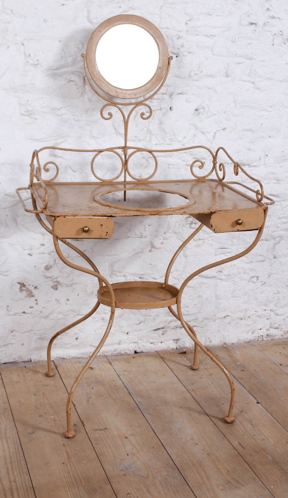 A painted beige wrought iron washstand featuring a top circular basin aperture, side towel rails, small drawers, and an attached swivel mirror with scrollwork.