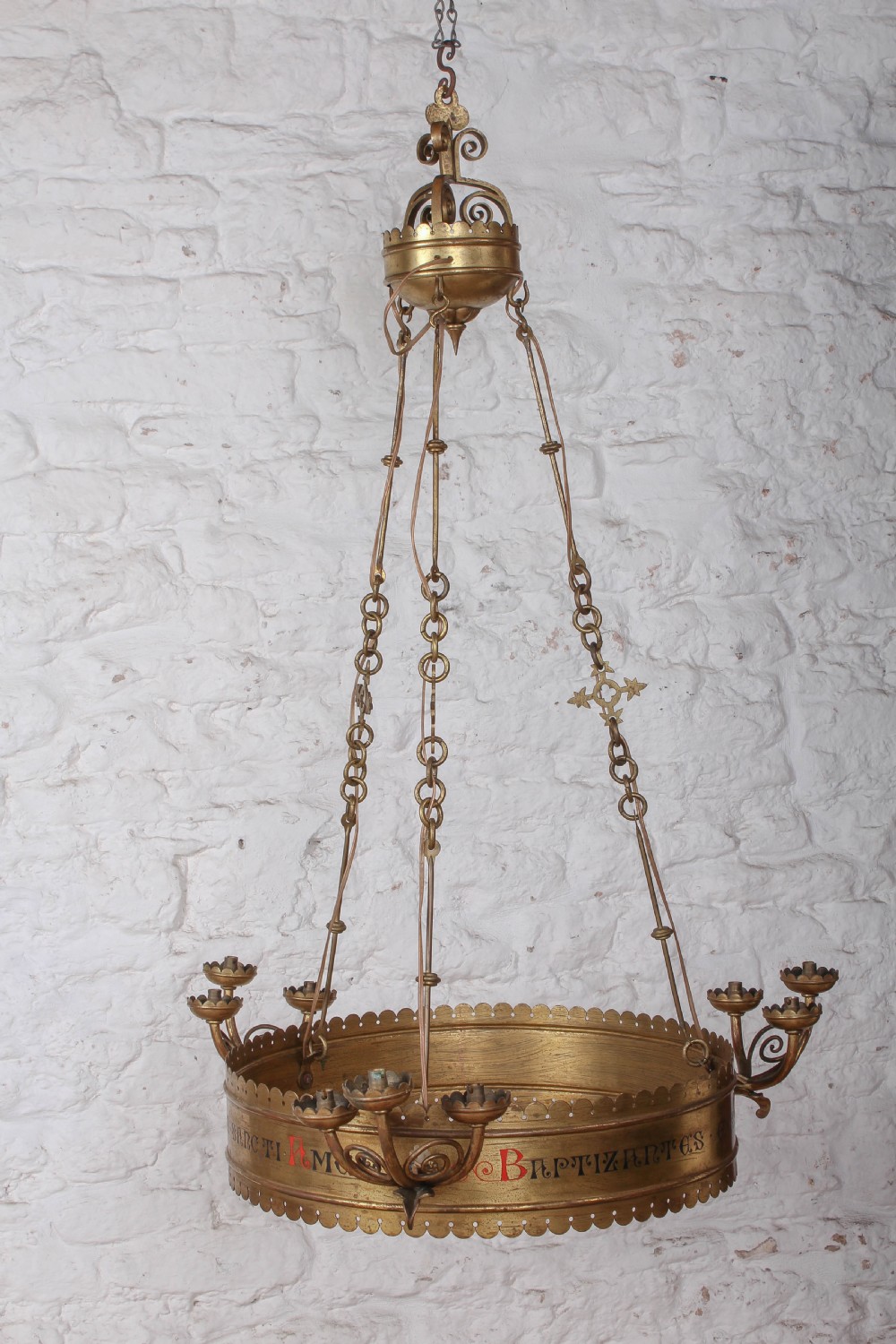 An Aesthetic Movement brass ecclesiastical corona chandelier featuring a circular band with painted Latin script, scalloped edges, multiple candle sconces, and hanging chains.