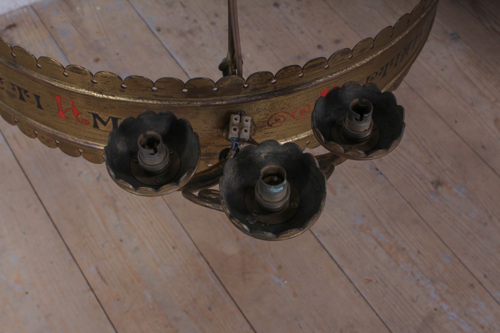 An Aesthetic Movement brass ecclesiastical corona chandelier featuring a circular band with painted Latin script, scalloped edges, multiple candle sconces, and hanging chains. View III