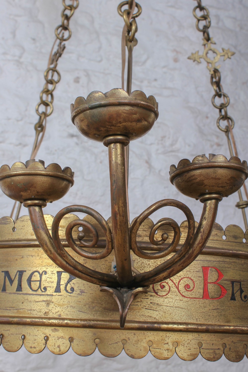 An Aesthetic Movement brass ecclesiastical corona chandelier featuring a circular band with painted Latin script, scalloped edges, multiple candle sconces, and hanging chains. View IV