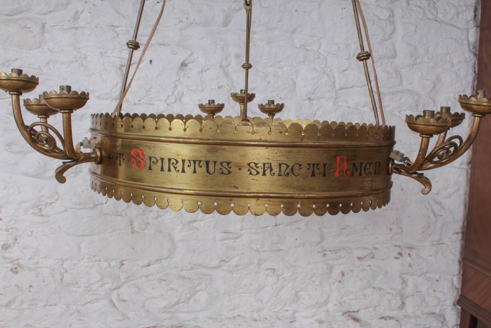 An Aesthetic Movement brass ecclesiastical corona chandelier featuring a circular band with painted Latin script, scalloped edges, multiple candle sconces, and hanging chains. View VII