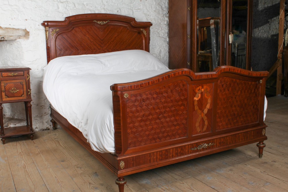An antique French mahogany bed frame featuring geometric parquetry panels, fruitwood ribbon marquetry on the footboard, and gilt bronze mounts.