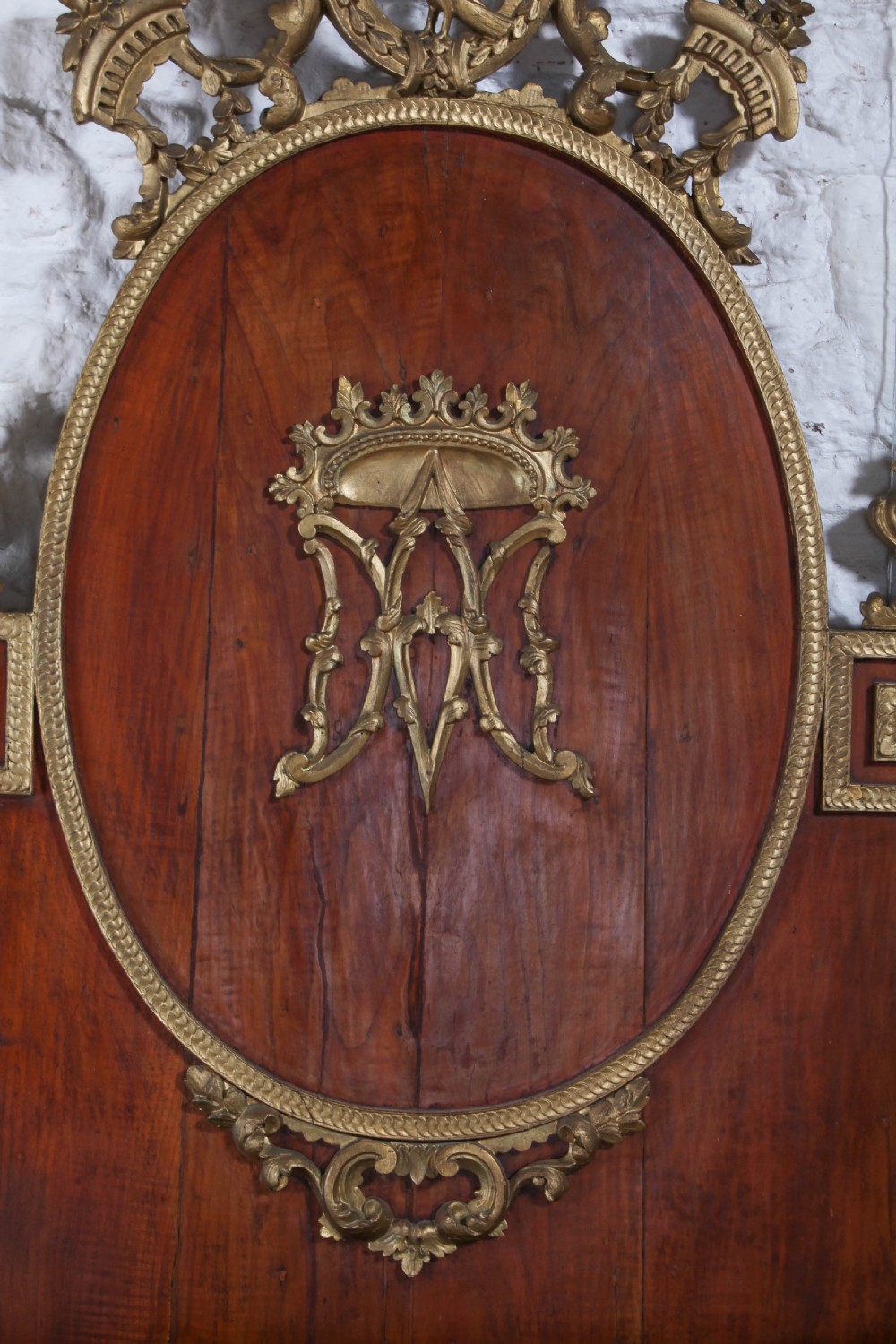 An antique mahogany super king bed featuring a high headboard with an oval central medallion, carved giltwood crest, Greek key motifs, and turned posts. View V