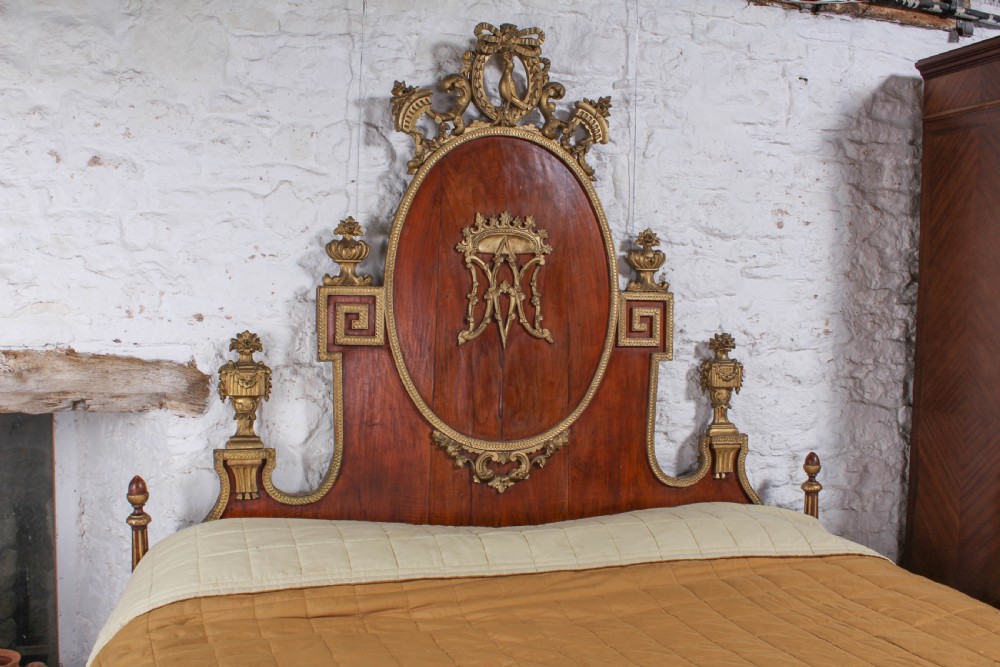 An antique mahogany super king bed featuring a high headboard with an oval central medallion, carved giltwood crest, Greek key motifs, and turned posts. View VI