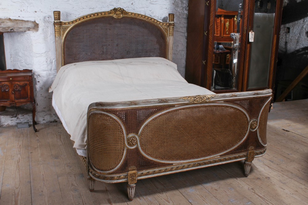 A Louis XVI style French corbeille bed featuring a carved giltwood frame, woven cane panels, fluted legs, and a rounded footboard.