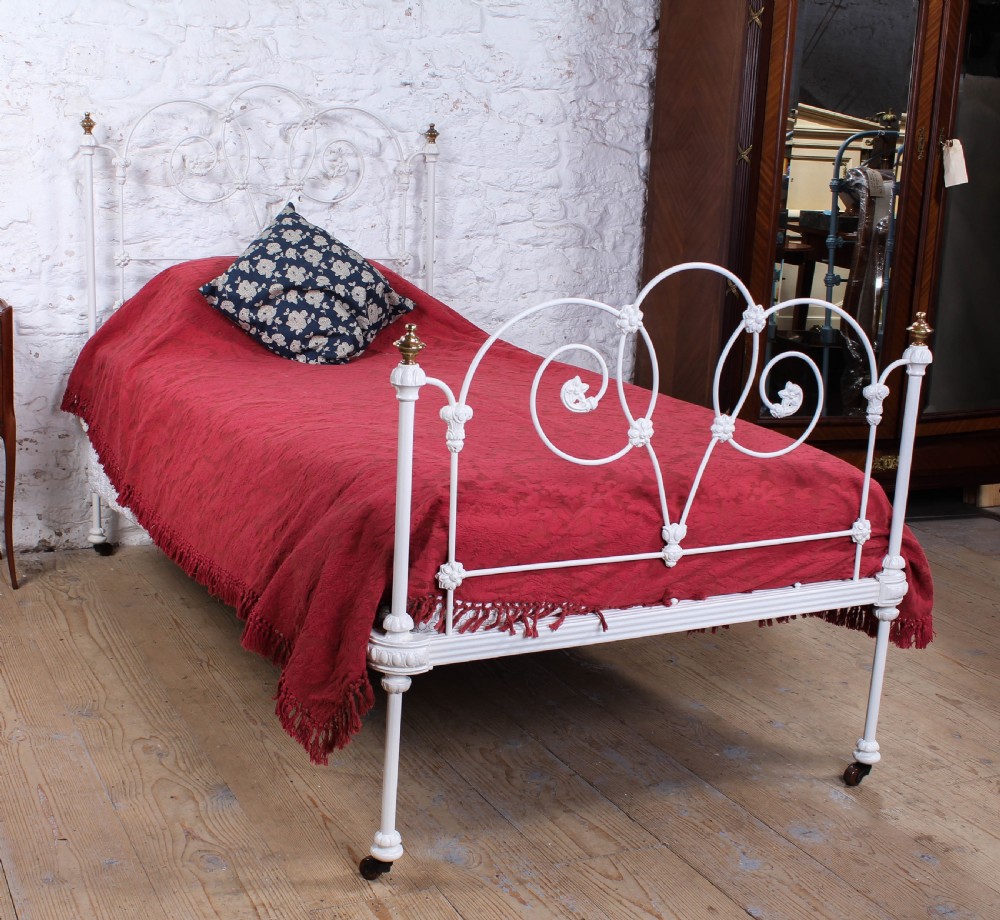 A Victorian white painted wrought iron single bed frame featuring ornate scrollwork, floral metal castings, turned brass finials, and castors.
