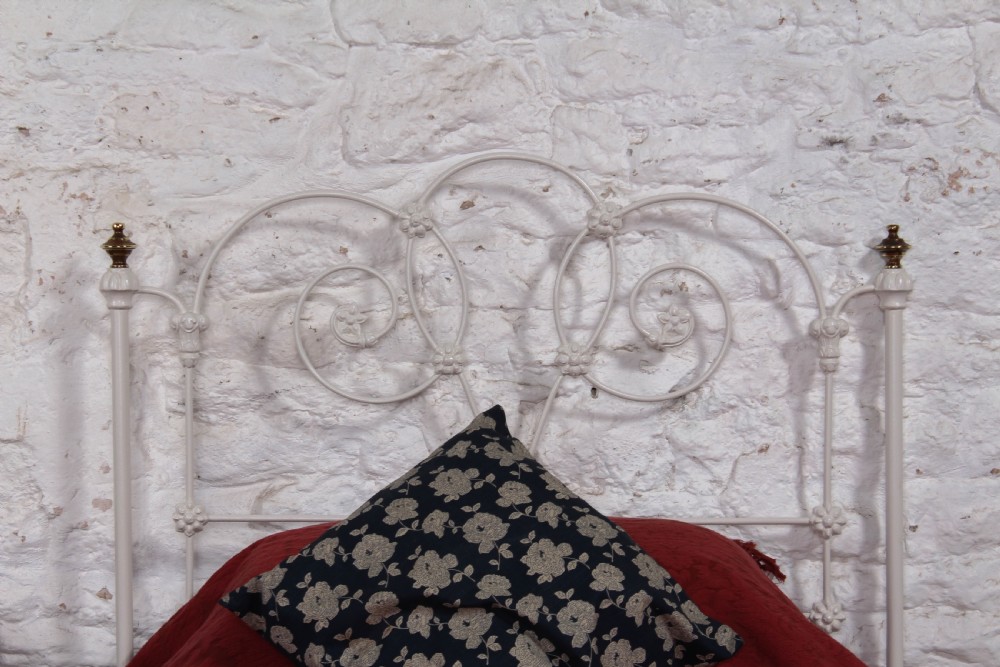 A Victorian white painted wrought iron single bed frame featuring ornate scrollwork, floral metal castings, turned brass finials, and castors. View V