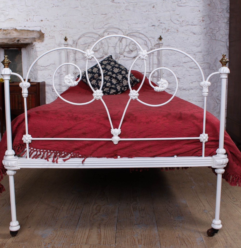 A Victorian white painted wrought iron single bed frame featuring ornate scrollwork, floral metal castings, turned brass finials, and castors. View VI
