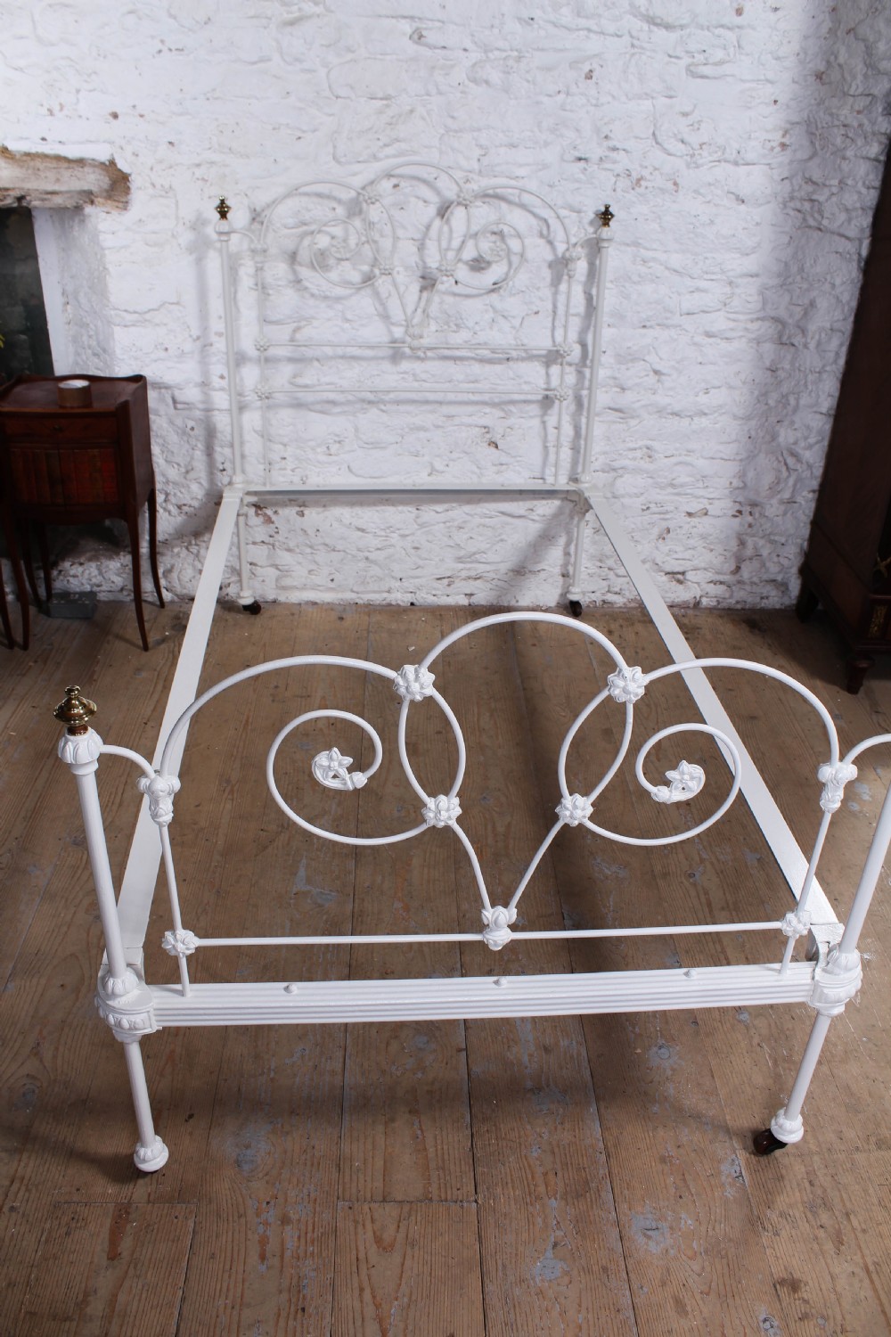 A Victorian white painted wrought iron single bed frame featuring ornate scrollwork, floral metal castings, turned brass finials, and castors. View IX