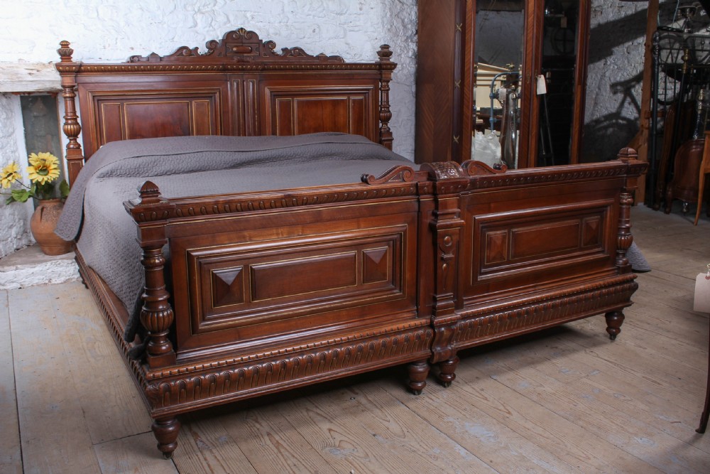 A carved solid walnut emperor size bedstead featuring a cresting motif, geometric panelled headboard and footboard, and turned posts.