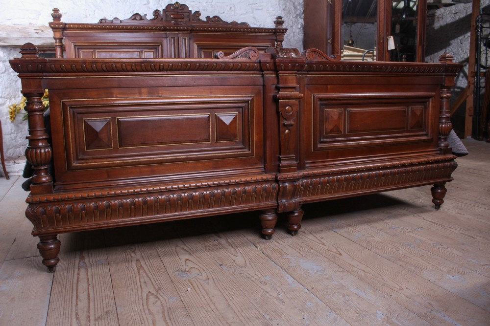 A carved solid walnut emperor size bedstead featuring a cresting motif, geometric panelled headboard and footboard, and turned posts. View III