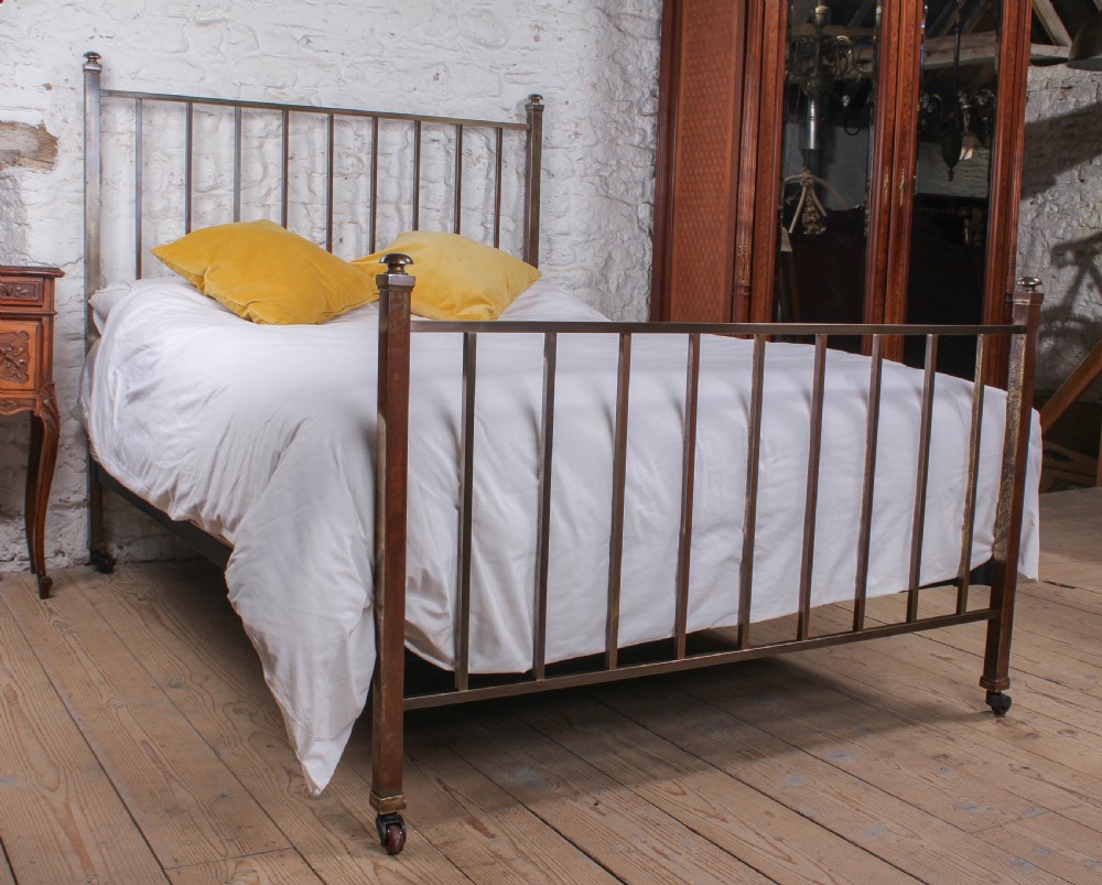 An antique Victorian king-size bed frame featuring vertical brass spindles, square corner posts, and an aged metallic patina set on castors.