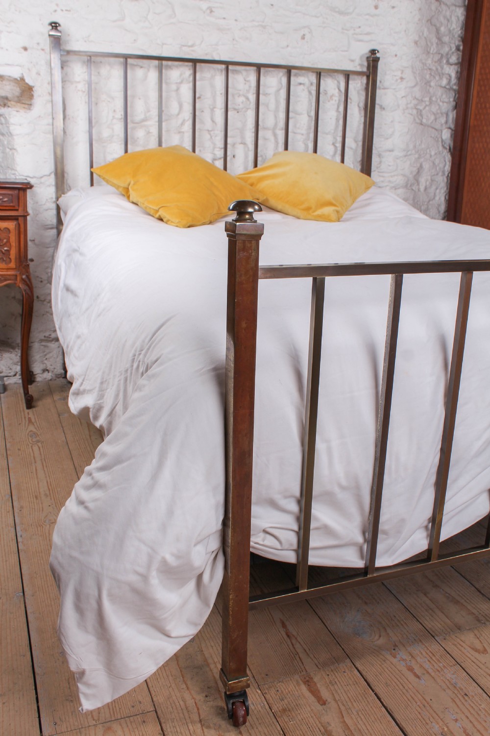 An antique Victorian king-size bed frame featuring vertical brass spindles, square corner posts, and an aged metallic patina set on castors. View VI