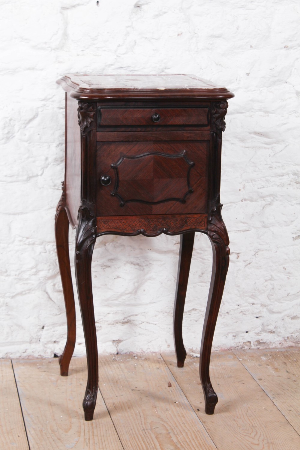 A dark wood Louis XV style bedside cabinet featuring an inset marble top, quarter-veneered door, drawer, carved apron, and tall cabriole legs.