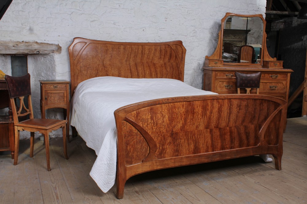 An Art Nouveau wooden bedroom suite featuring a curved double bedstead, matching dressing table with mirror, nightstand, and leather-upholstered side chairs.