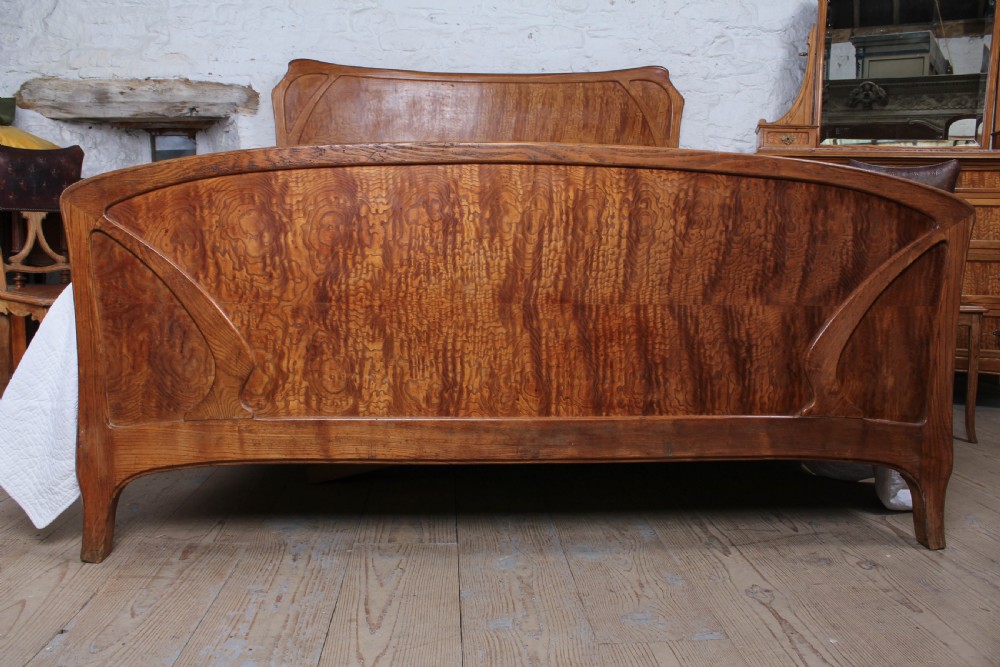 An Art Nouveau wooden bedroom suite featuring a curved double bedstead, matching dressing table with mirror, nightstand, and leather-upholstered side chairs. View II