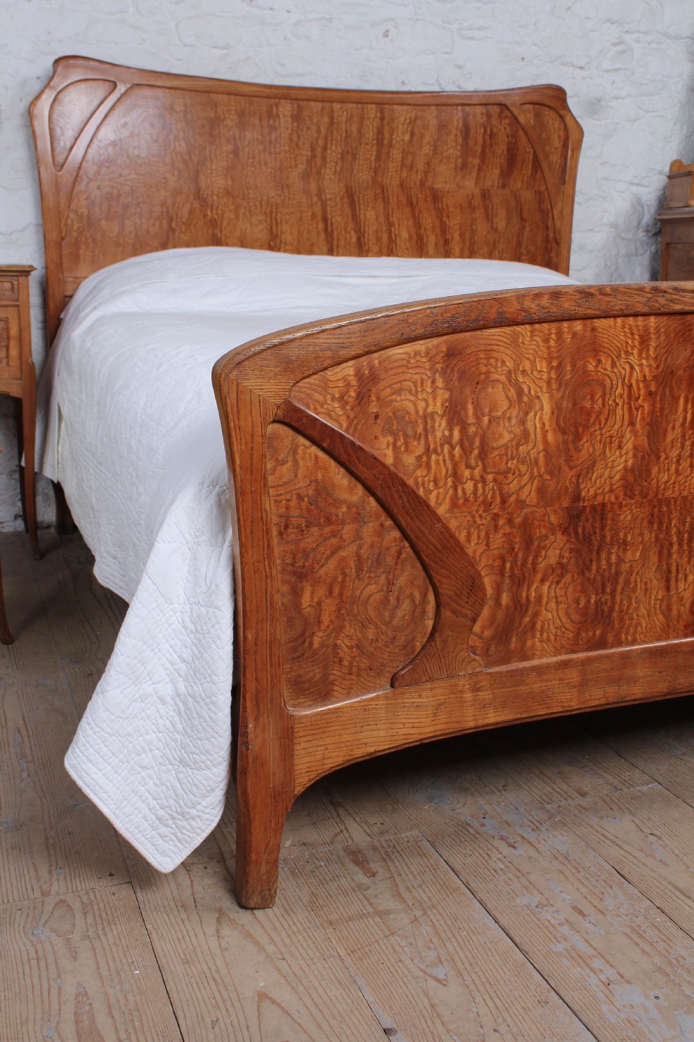 An Art Nouveau wooden bedroom suite featuring a curved double bedstead, matching dressing table with mirror, nightstand, and leather-upholstered side chairs. View VI