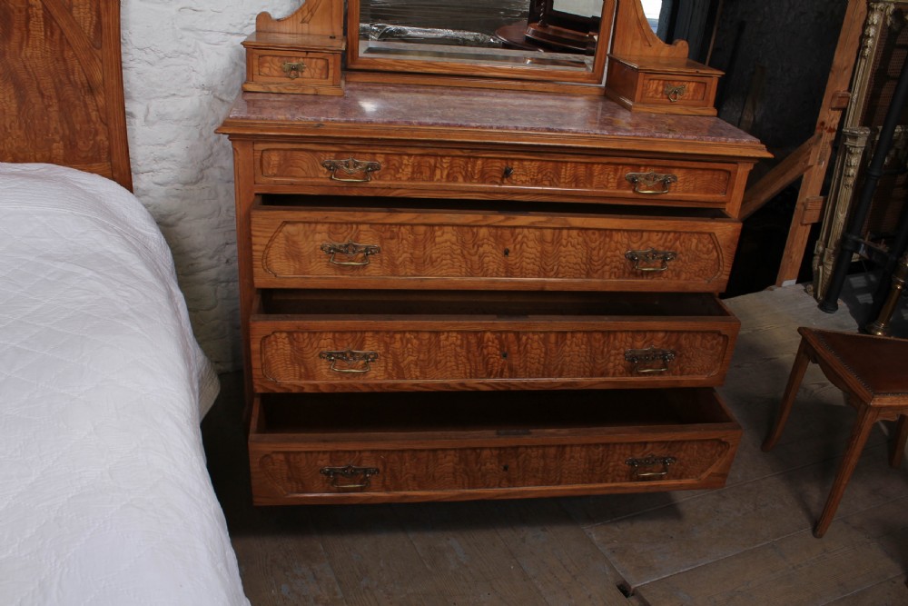 An Art Nouveau wooden bedroom suite featuring a curved double bedstead, matching dressing table with mirror, nightstand, and leather-upholstered side chairs. View XV