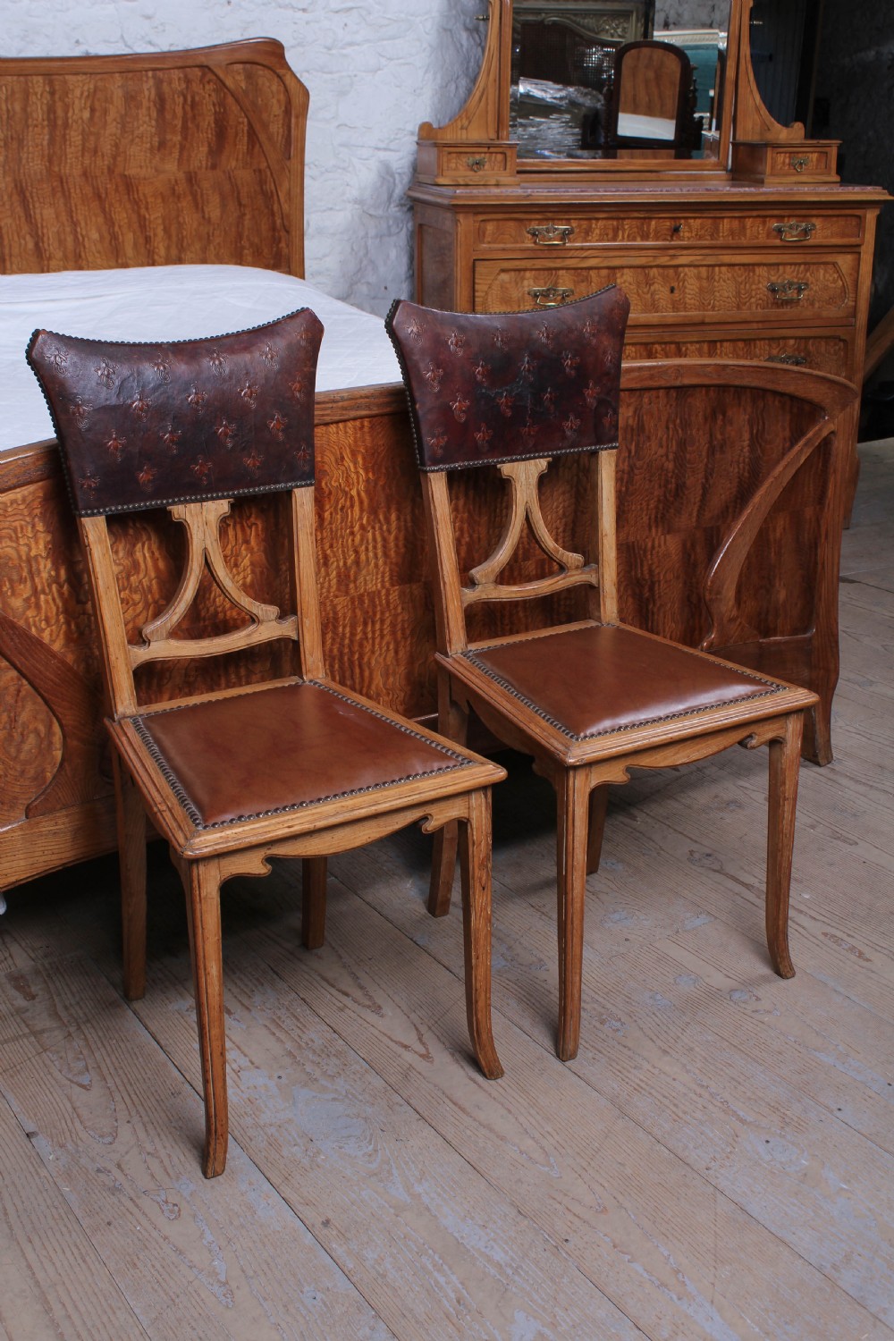 An Art Nouveau wooden bedroom suite featuring a curved double bedstead, matching dressing table with mirror, nightstand, and leather-upholstered side chairs. View XVI