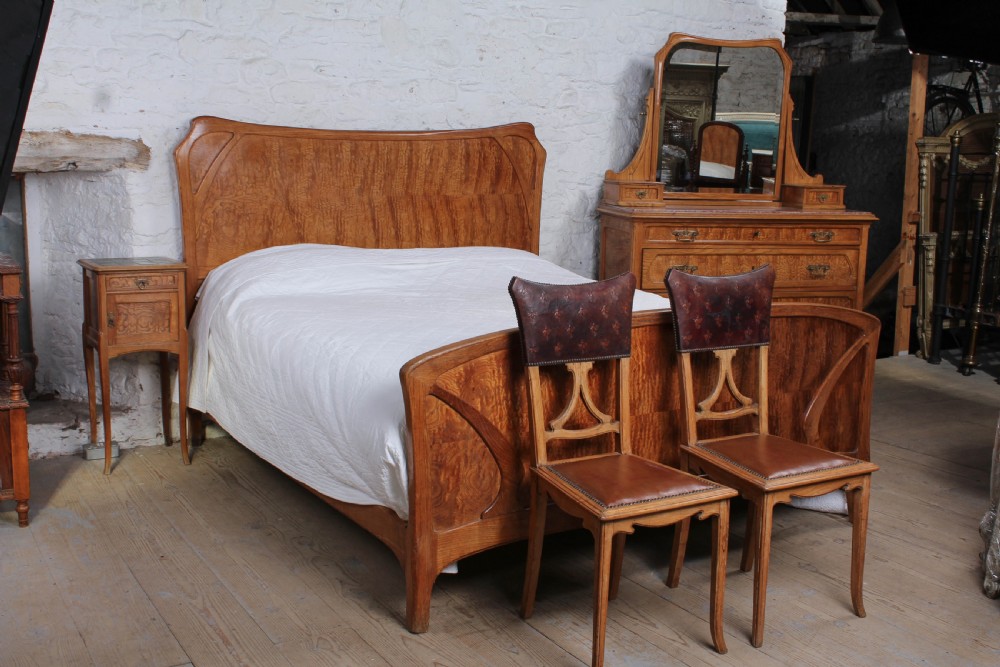 An Art Nouveau wooden bedroom suite featuring a curved double bedstead, matching dressing table with mirror, nightstand, and leather-upholstered side chairs. View XX