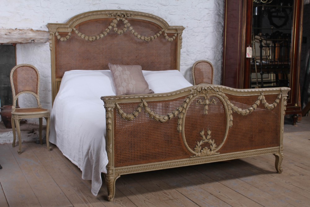 A French Louis XVI style super king bedstead featuring woven rattan cane panels, carved floral swags, and matching caned side chairs in a painted distressed finish.