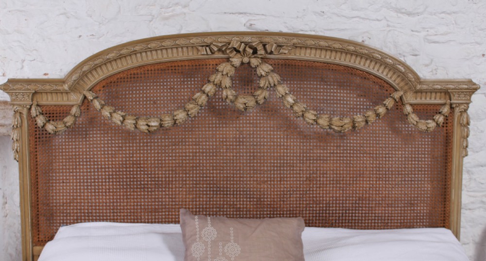 A French Louis XVI style super king bedstead featuring woven rattan cane panels, carved floral swags, and matching caned side chairs in a painted distressed finish. View VI