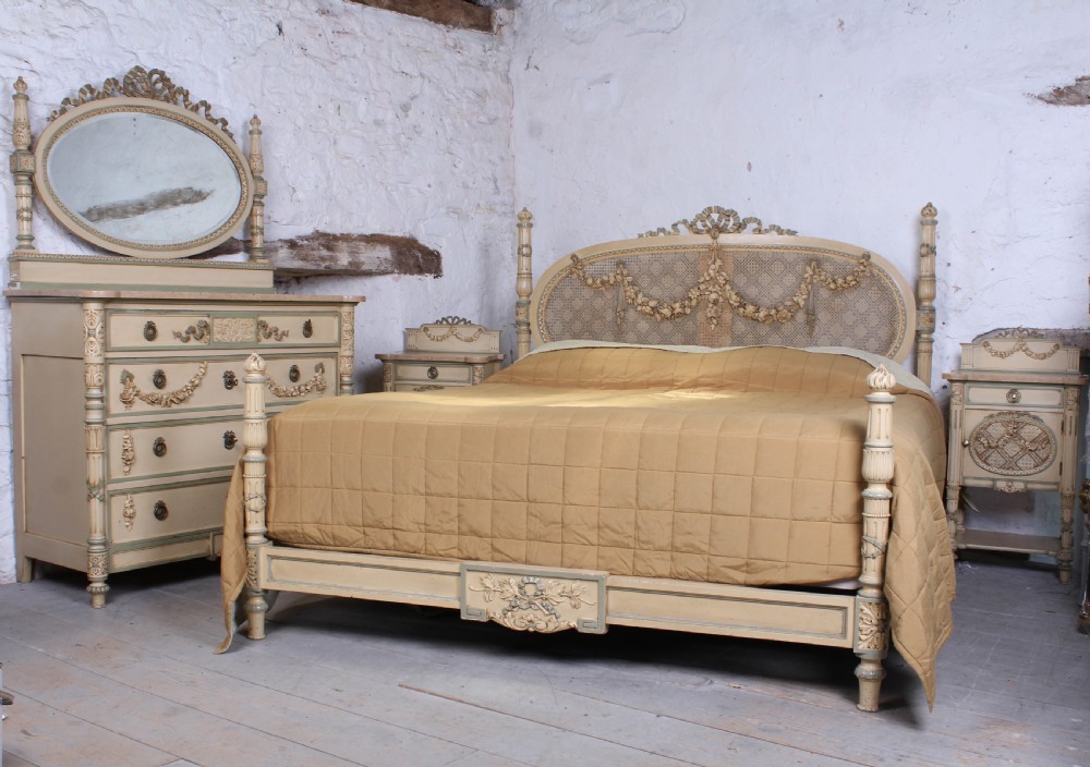 A cream and green painted super king size bedroom set featuring a detailed caned headboard, carved garlands, two matching bedside tables, and a chest of drawers with an oval mirror.