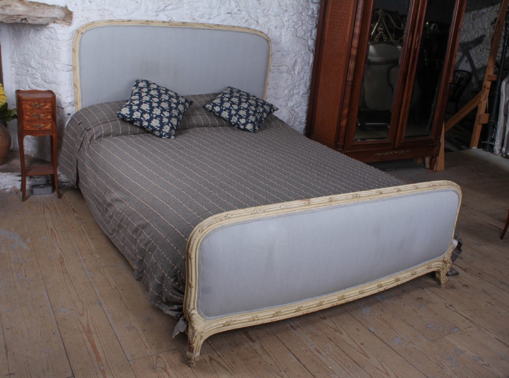 An antique French king-size bed featuring a cream painted carved wooden frame and light grey upholstered panels on the headboard and footboard.