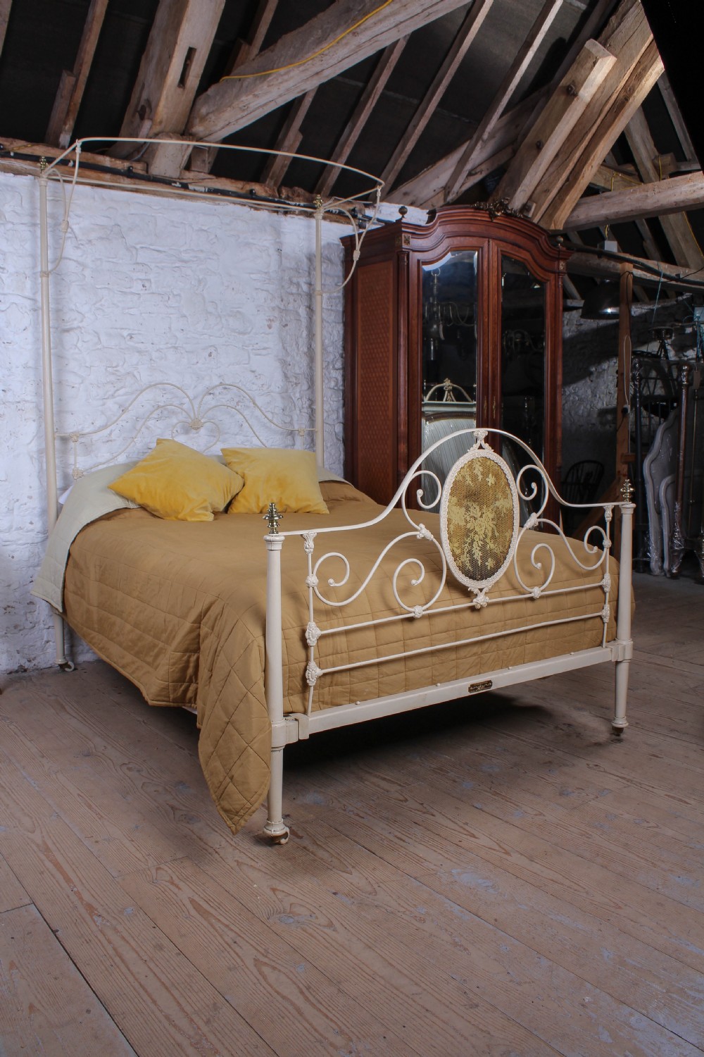A cream painted wrought iron king size half tester bed frame featuring an ornate scrollwork footboard with a central oval medallion and brass finials.