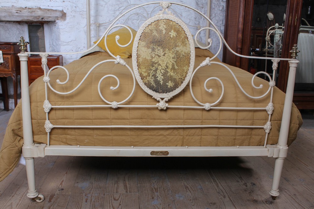 A cream painted wrought iron king size half tester bed frame featuring an ornate scrollwork footboard with a central oval medallion and brass finials. View II