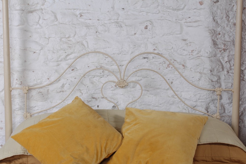 A cream painted wrought iron king size half tester bed frame featuring an ornate scrollwork footboard with a central oval medallion and brass finials. View VI