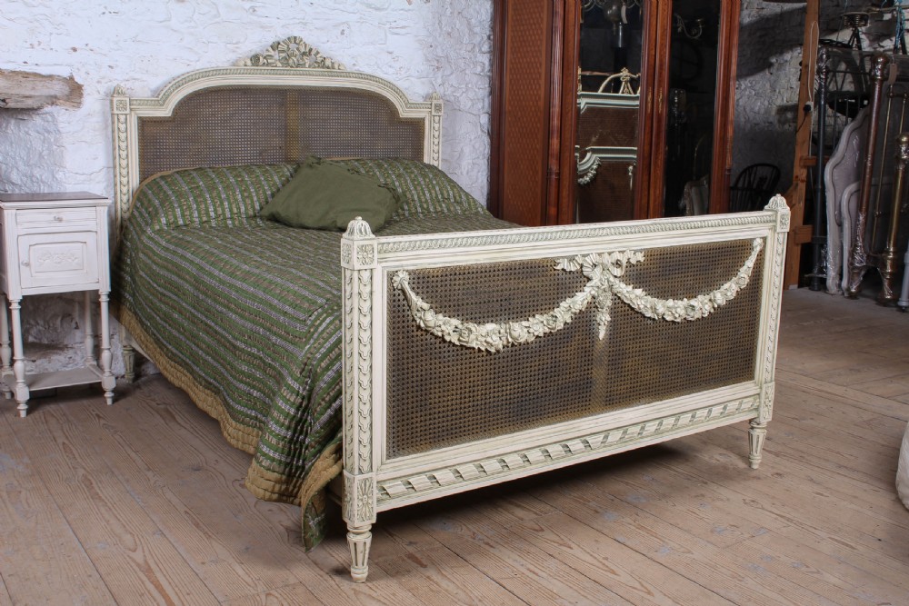 A painted Louis XVI style double bed frame featuring carved floral garlands, fluted corner posts, and woven cane panels in the headboard and footboard.
