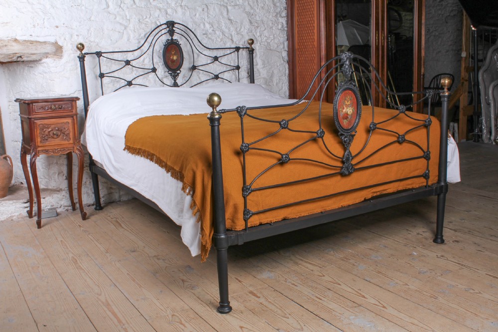 A Victorian black-painted cast iron bedstead featuring brass finials, arched lattice scrollwork, and oval floral enamel medallions on the headboard and footboard.