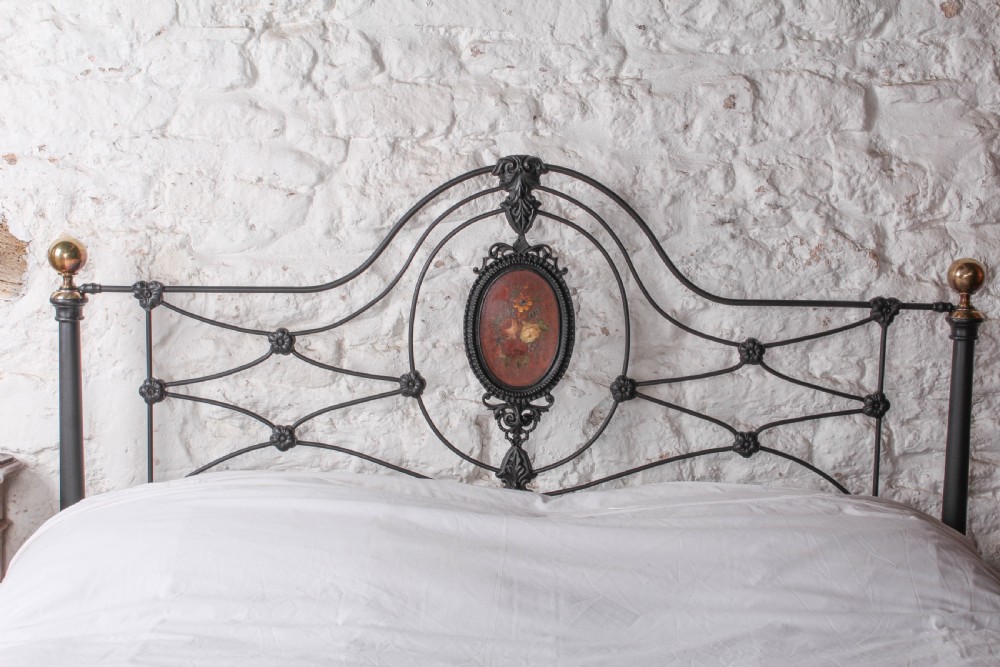 A Victorian black-painted cast iron bedstead featuring brass finials, arched lattice scrollwork, and oval floral enamel medallions on the headboard and footboard. View V